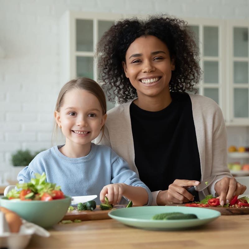 Portrait of smiling African American mom and little Caucasian daughter look at camera cooking in kitchen together, happy biracial mother and small girl child have fun preparing healthy food at home